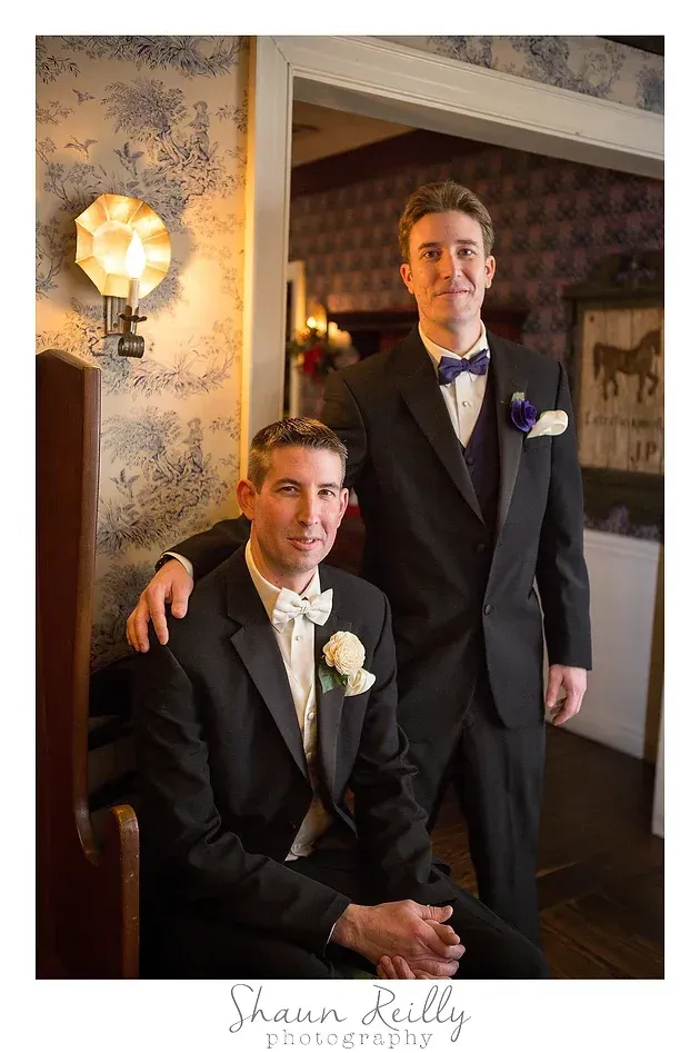 Two men in tuxedos posing indoors beside a patterned wall and lamp