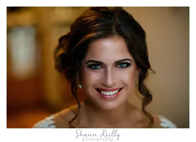 Smiling woman with dark hair and makeup, facing the camera against a warm, blurred background