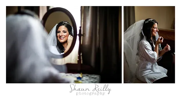 Bride smiling in mirror while getting ready in a white robe and veil near a window