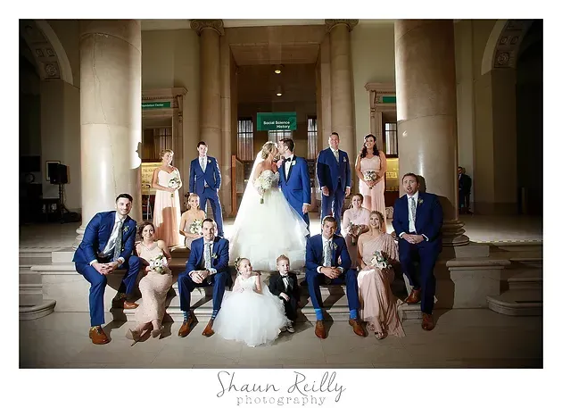 Wedding party posed on steps in a grand stone entryway, with bride and groom centered under columns