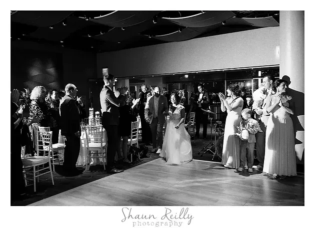 Wedding reception guests cheer as a bride and groom dance in a black-and-white ballroom photo