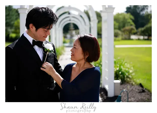 Man in black tuxedo and woman in navy dress share a tender look in a bright garden archway.