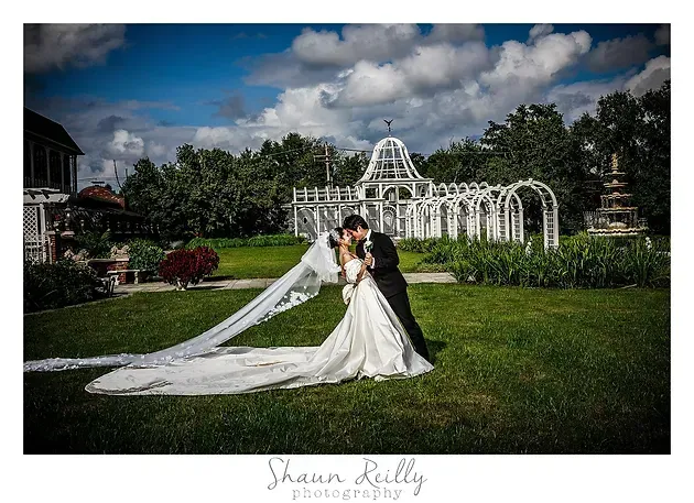 Bride and groom posing on a lawn before a white garden arbor, with a long veil and train in sunlight