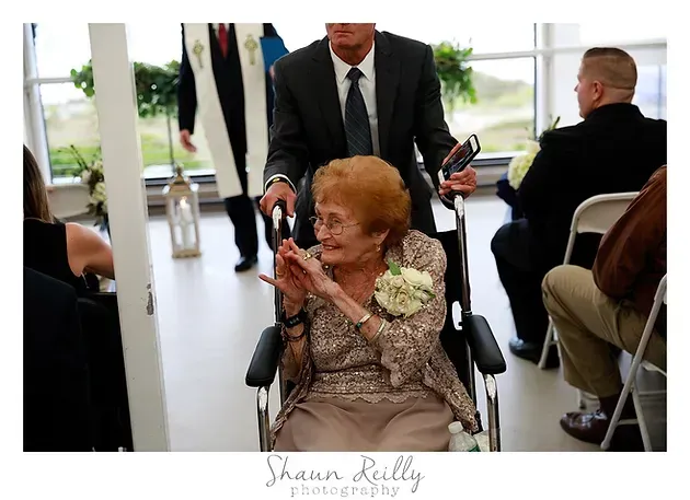 Elderly woman in a wheelchair clapping at a formal indoor event, with a suited man standing behind her