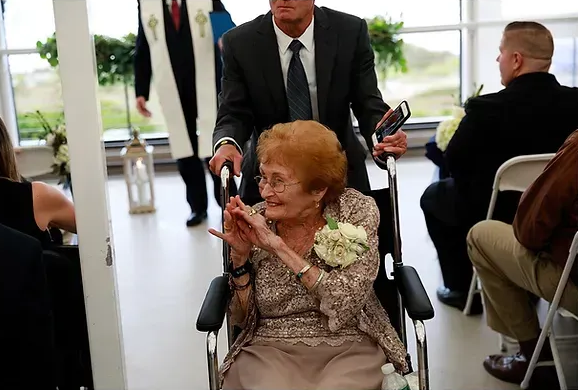 Woman in a beige dress claps while seated in a wheelchair at an indoor event, with a man standing behind her.