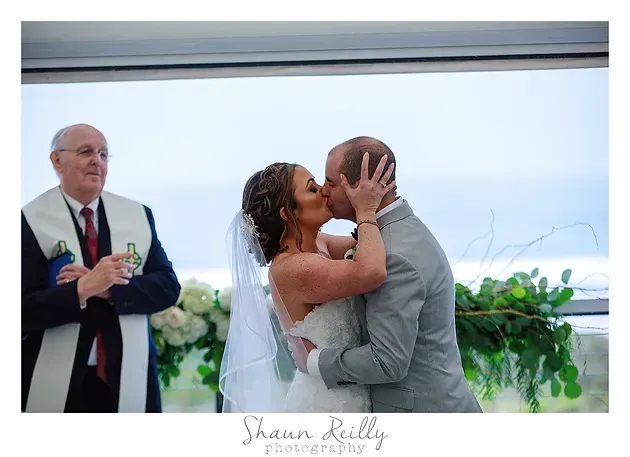 Bride and groom kiss at wedding ceremony as officiant looks on