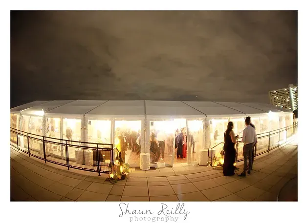 Nighttime wedding reception under a white tent with guests standing near warm lights and railing