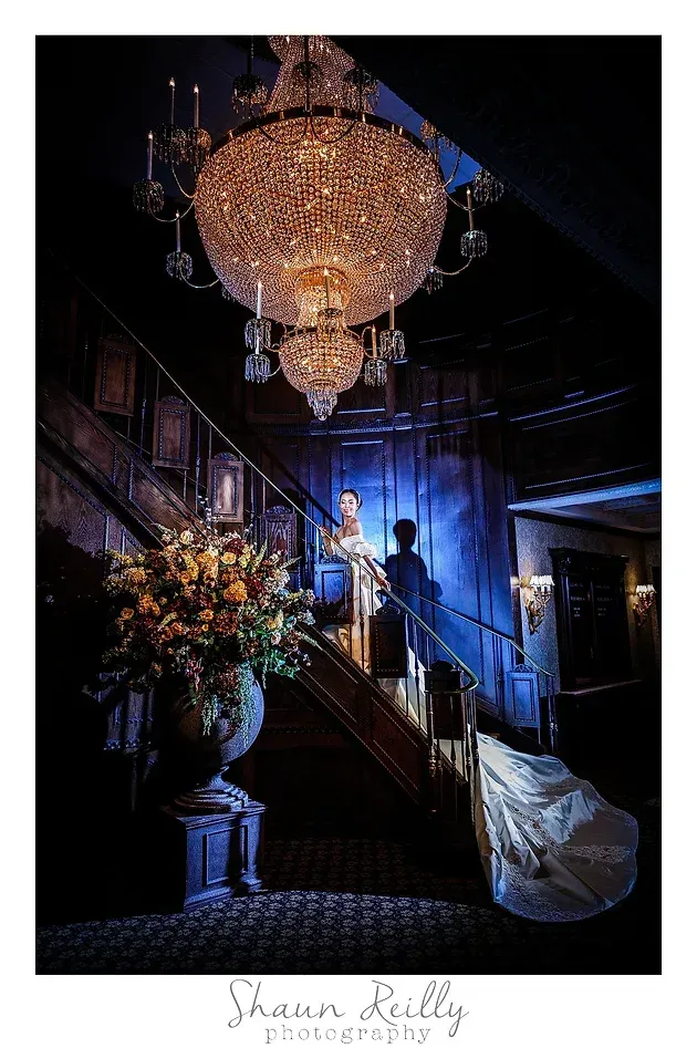 Elegant chandelier-lit staircase in a dim hall with flowers and a person in a blue dress descending