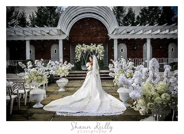 Bride in long white gown standing in a floral wedding arch and garden aisle, surrounded by white flowers