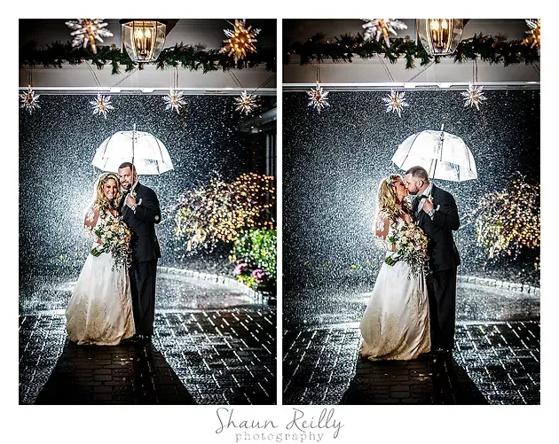 Newlyweds kiss under an umbrella in the rain at a lit outdoor wedding entrance.