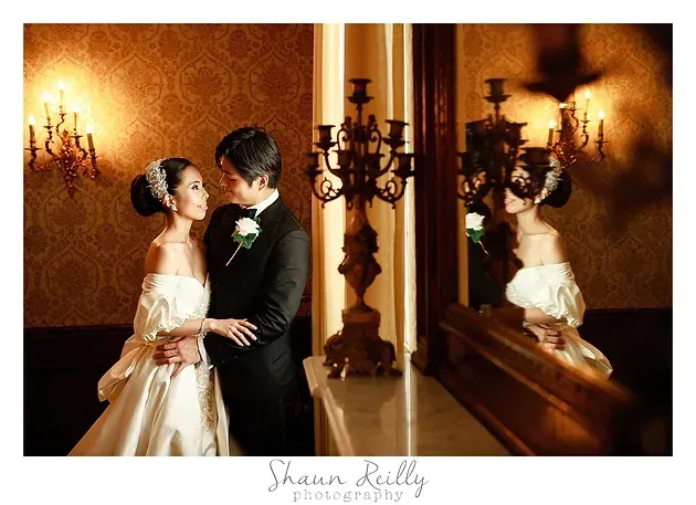 Bride and groom embrace in a warmly lit ballroom with chandeliers and mirrored wall.