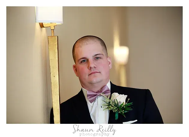 Man in a tuxedo with a pink bow tie and boutonniere standing indoors beside a lamp