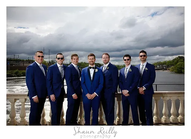 Seven men in blue suits standing by a river under a cloudy sky