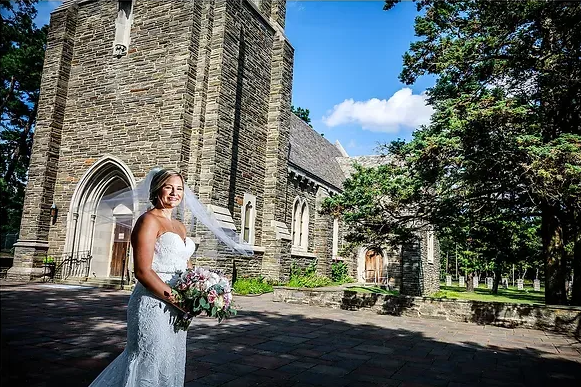 Bride in a white dress holding a bouquet beside a stone church on a sunny day