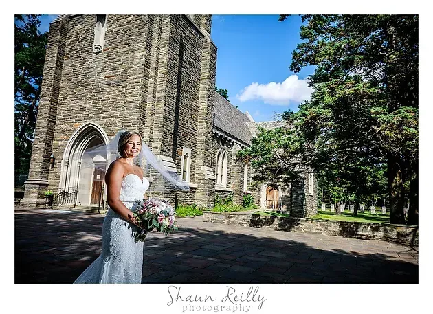 Bride in a white dress holding a bouquet outside a stone church on a sunny day