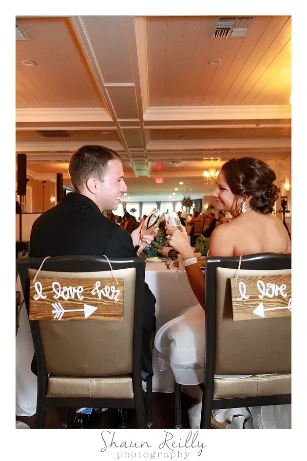 Guests seated at a reception table, facing each other and holding hands in a warmly lit hall