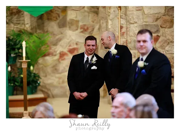 Three men in black suits stand at a wedding ceremony beside a stone wall and candlelit altar