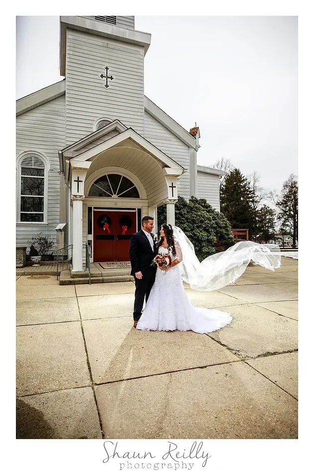 Wedding couple posing outside a church, bride’s veil blowing in the wind