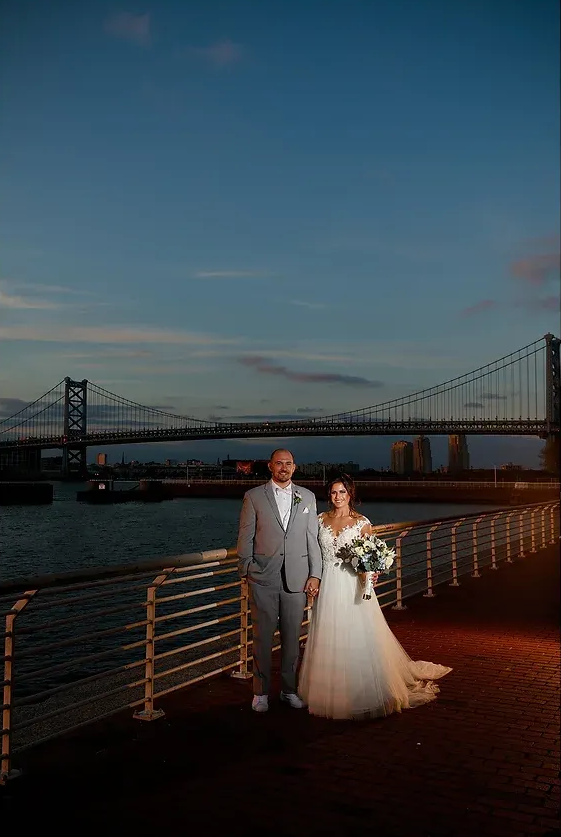 Bride and groom pose on a waterfront deck at sunset with a suspension bridge in the background.