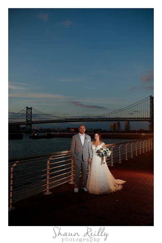 Bride and groom in wedding attire on a waterfront pier at sunset with a bridge in the background