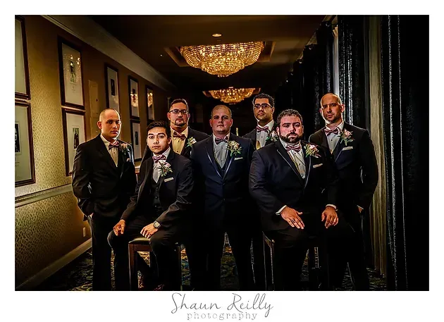Six men in tuxedos pose in a dimly lit hallway with a chandelier overhead.