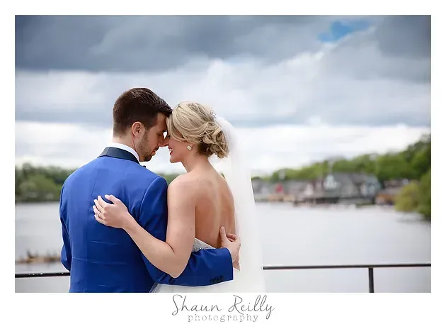 Couple embracing by a waterfront, with cloudy sky and blurred shoreline in the background.