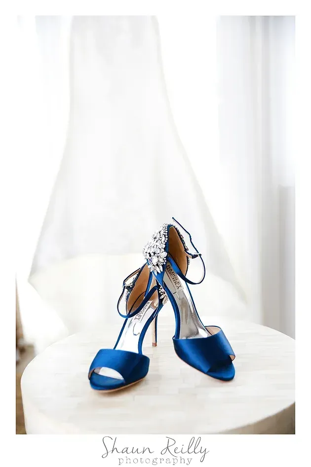 Blue satin high heels with jeweled straps on a white surface near a sheer curtain