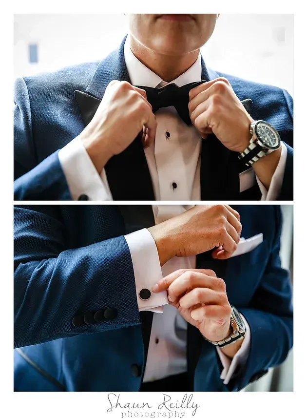 Man in a blue suit adjusting a black bow tie and cufflinks on a white shirt.
