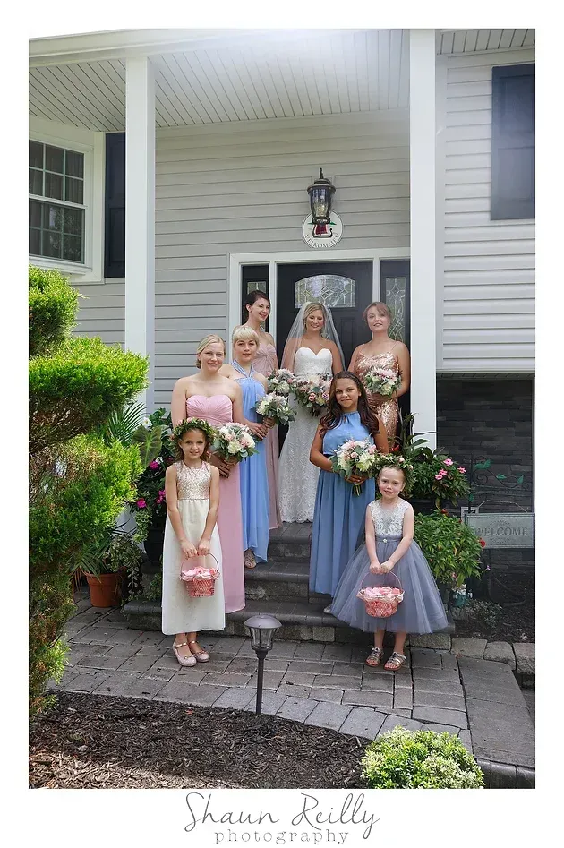 Group of children and adults posing on a porch, holding bouquets and wearing pastel dresses and suits.