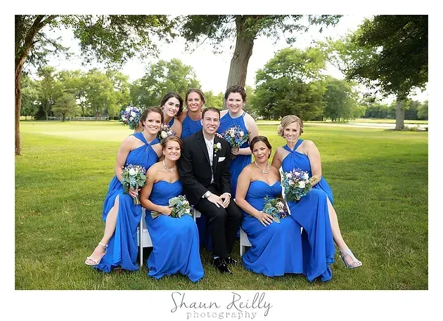 Wedding party in blue dresses and black suits posing on a lawn with trees behind them