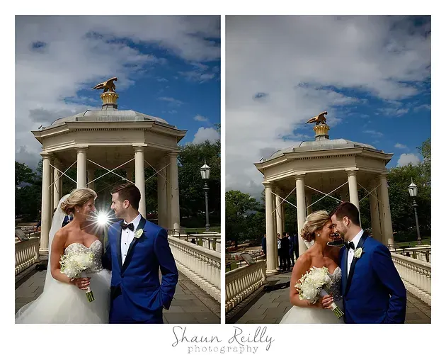 Bride and groom pose in blue suits and white dress under a gazebo with a gold statue.