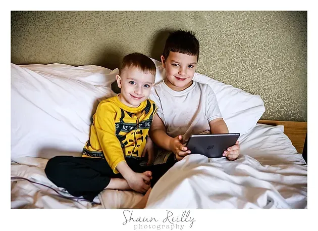 Two children on a bed looking at a tablet, one in a yellow shirt and one in white, smiling