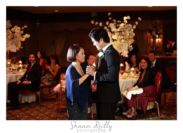 Couple dancing at a wedding reception, holding hands amid seated guests and warm floral décor