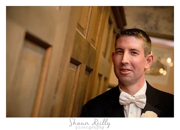 Man in a tuxedo with a white bow tie standing by a wooden wall, smiling softly.