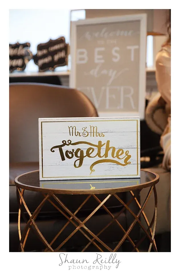 Gold and white “Let’s Stay Together” sign on a round wire table with a blurred “best day ever” sign behind.