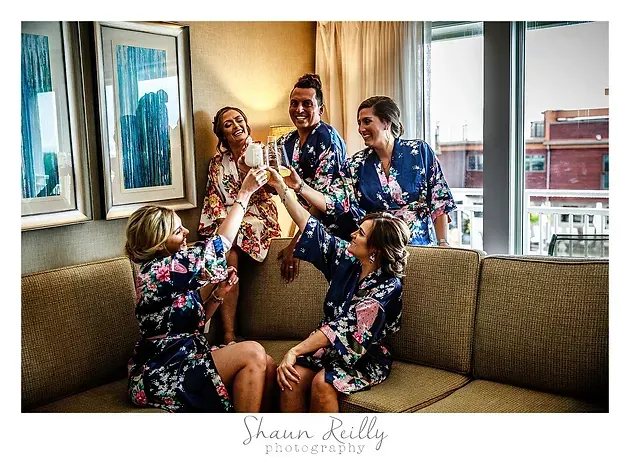 Four people in floral robes laugh and toast with champagne in a bright living room by a window.