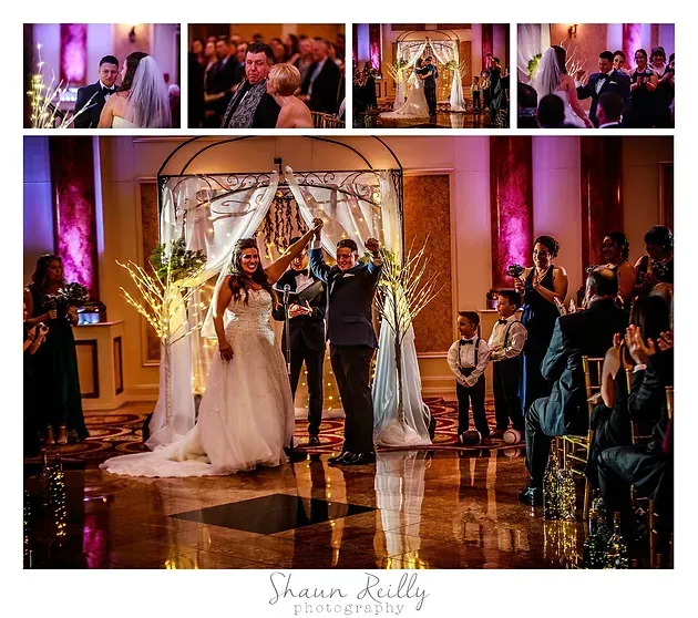 Wedding couple under a floral arch as guests applaud in a warmly lit ballroom