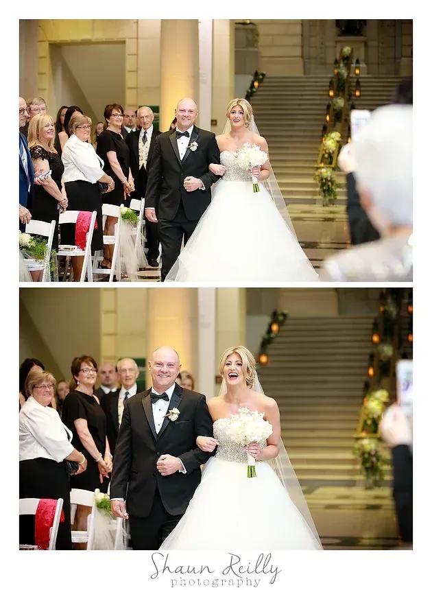 Bride walks down church aisle with escort, guests watching, staircase and floral decorations behind them.