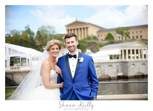 Wedding couple posing by a waterfront in front of a classical building, groom in blue suit, bride in white dress