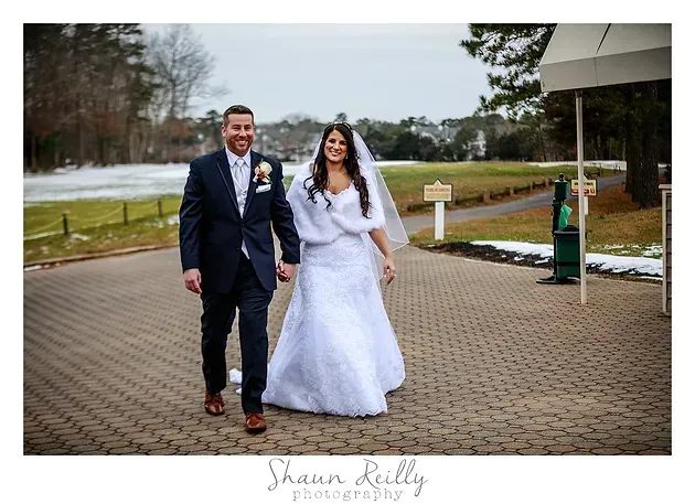 Couple in wedding attire walking on a paved path near a snowy lakeside park