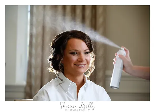 Woman smiles as hairspray is applied to her styled updo in a softly lit room.