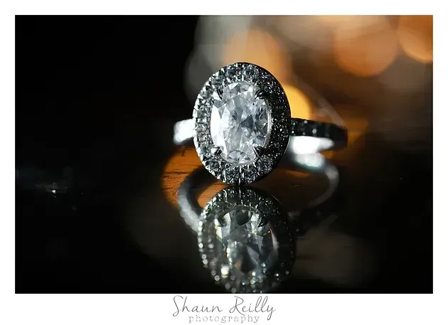 Oval diamond halo ring on a reflective black surface with warm bokeh background