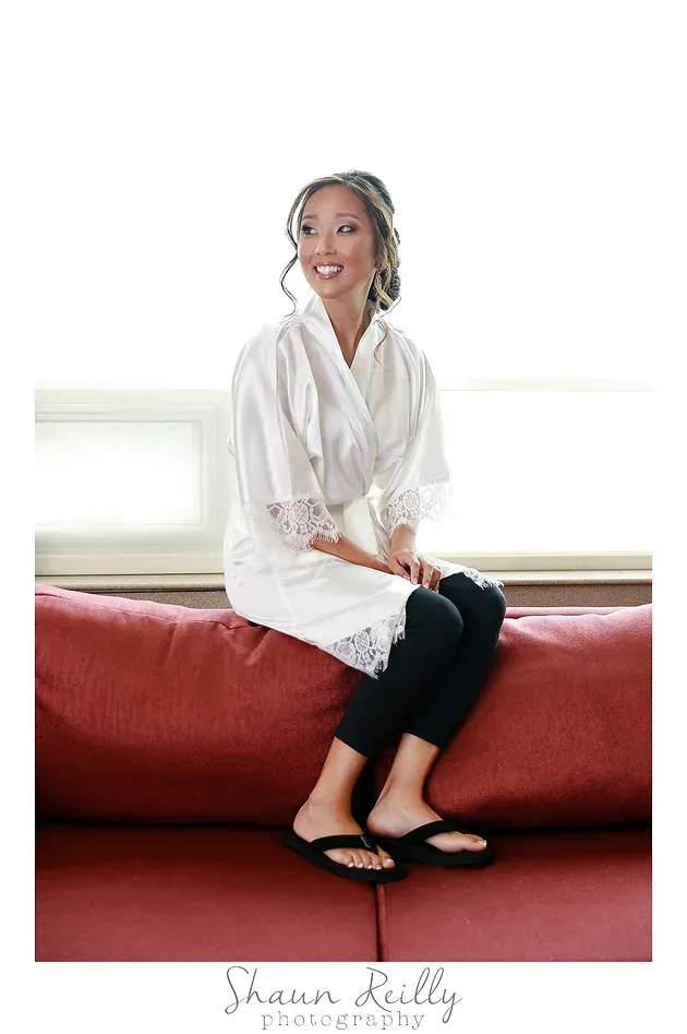 Woman in white blouse and black pants sitting on a red bed, smiling in a bright room.