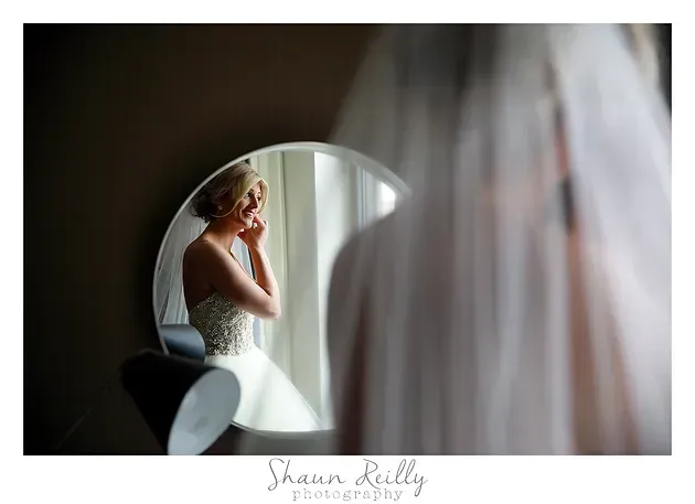 Bride adjusting earring in mirror, white dress and veil, soft window light