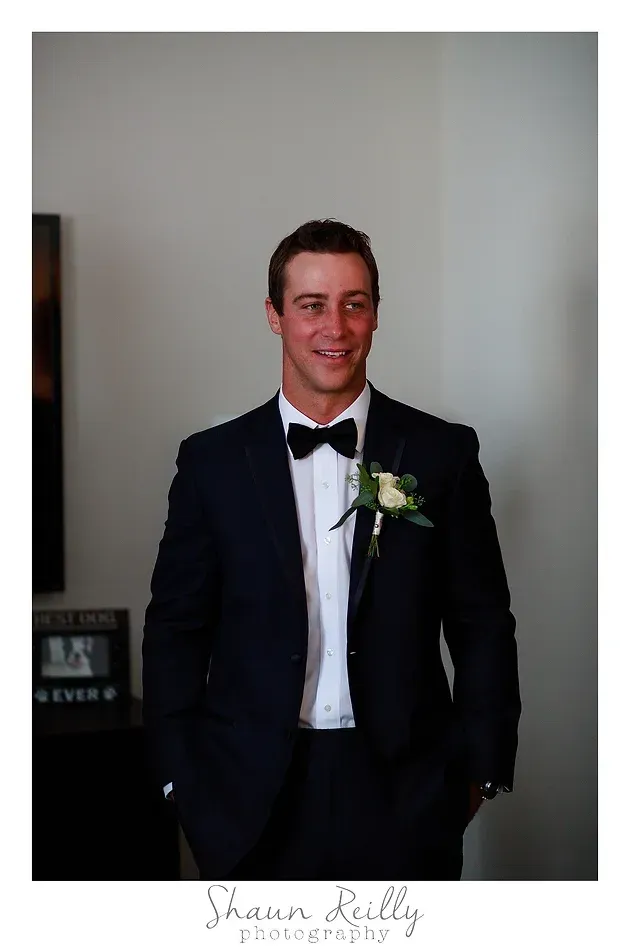 Man in a black tuxedo with a white boutonniere, smiling indoors against a plain wall