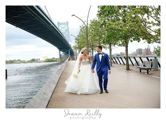 Bride and groom walking hand in hand by a river under a bridge on a cloudy day