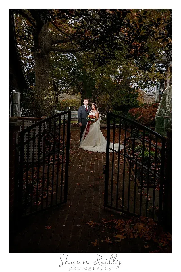Bride and groom standing on a tree-lined path behind open iron gates in an autumn garden