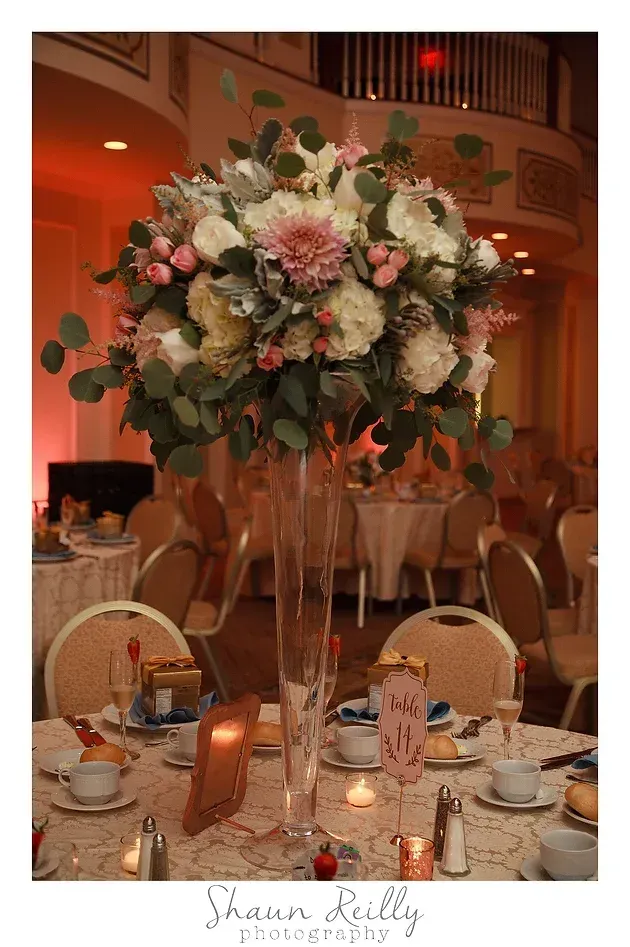 Elegant wedding reception table with tall pink-and-white floral centerpiece in a warm orange-lit venue