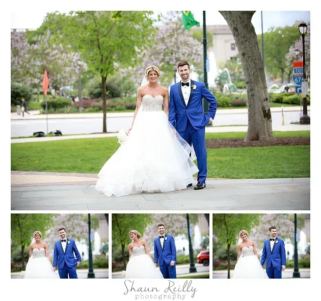 Bride and groom in a blue suit and white gown posing outdoors in a park with trees and grass.