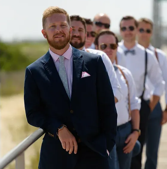 Group of people in formal attire outdoors, led by a man leaning on a railing in a navy suit.